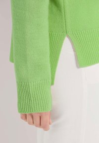 Grüner gestrickter Pullover mit gerippten Bündchen und seitlichen Schlitzen, kombiniert mit weißen Hosen. Die Textur ist glatt und leicht elastisch.