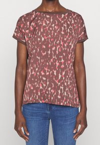 Persona vistiendo una camisa de manga corta marrón con un patrón abstracto en rosa y blanco, y jeans azules, de pie frente a un fondo liso.