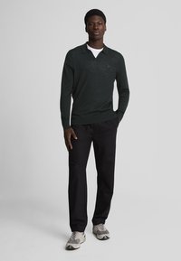 Dunkelgrüner Poloshirt-Pullover mit langen Ärmeln und V-Ausschnitt, kombiniert mit schwarzen Hosen und hellen Sneakers. Minimalistisches Design, strukturierter Stoff.