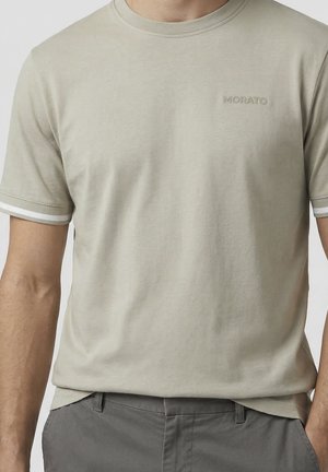 Homme portant un T-shirt beige à manches courtes avec un bord blanc sur les manches et le logo "MORATO" sur la poitrine, assorti à un pantalon gris.