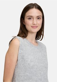 Ärmelloser, hellgrauer Strickpullover mit Rundhalsausschnitt, der ein strukturiertes Muster und fransige Kanten an den Armöffnungen aufweist.