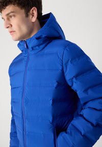 Chaqueta acolchada azul con cuello alto y capucha, que cuenta con un cierre frontal de cremallera y paneles cosidos horizontalmente. El material parece brillante.
