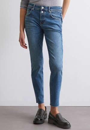 Kvinde iført blå jeans med høj talje og smal pasform med frynset kant samt sorte læder loafers, stående mod en simpel lys væg.