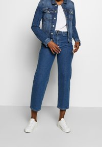 Jeansjacka med knäppning framtill, två bröstfickor och krage i klassisk stil, kombinerad med högmidjade, vida jeans i mörkblått. Vita sneakers.