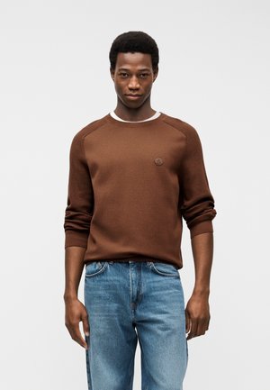 Ung mand iført en brun sweater over en hvid skjorte og blå jeans, stående mod en ensartet lysegrå baggrund.