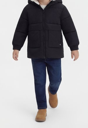 Enfant portant une doudoune noire, un jean bleu foncé et des bottes couleur camel marchant vers l'avant sur un fond blanc.