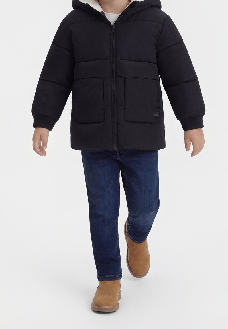 Enfant portant une doudoune noire, un jean bleu foncé et des bottes couleur camel marchant vers l'avant sur un fond blanc.