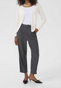 Nainen, jolla on valkoinen neuletakki valkoisen paidan päällä, tummanharmaat korkeavyötäröiset housut ja mustat kärjelliset kitten heel -mullit, seisoo yksi käsi taskussa.