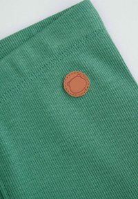 Zielony żeberkowany materiał z okrągłą brązową skórzaną łatką przedstawiającą wzór niedźwiedzia. Tekstura jest miękka, a brzegi mają szczegółowe przeszycia.