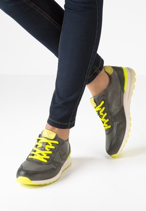 Persona con jeans azul oscuro y zapatillas grises con cordones y suelas de color amarillo brillante, de pie sobre una superficie blanca.