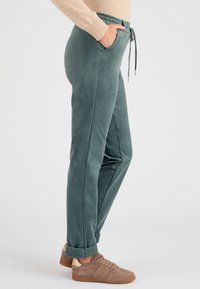 Pantaloni in corduroy verde con una vestibilità rilassata, tasche laterali, vita con coulisse e orli arrotolati, abbinati a sneaker color marrone chiaro.