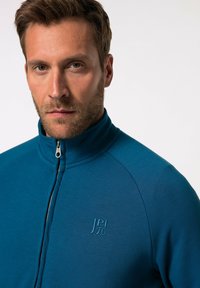 Chaqueta azul con cremallera, hecha de una tela suave, con cuello alto y un logo en relieve en el pecho. Textura suave con un diseño ajustado.