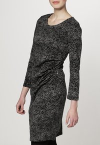 Robe à motifs noir et blanc avec un encolure dégagée, des manches longues et une silhouette ajustée mettant en valeur un tissu froncé sur le côté.