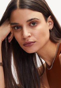 Primo piano di una donna con lunghi capelli scuri, che indossa una collana in argento e un piccolo orecchino a cerchio, con un colore di rossetto marrone opaco.