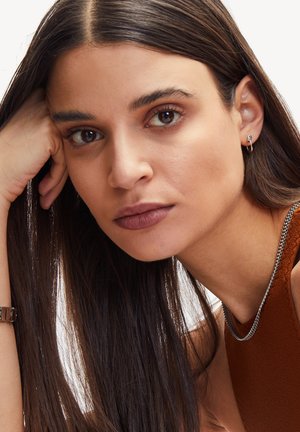 Gros plan sur une femme aux cheveux longs et foncés, portant un collier en chaîne argenté et une petite boucle d'oreille, avec une couleur de lèvres marron mate.