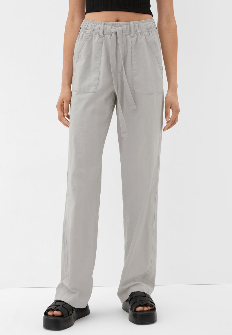 Femme portant un pantalon ample gris clair avec poches avant et taille à nouer, un top court noir et des sandales à plateforme noires sur fond blanc.
