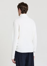 Maglione bianco a collo alto con costine, caratterizzato da maniche lunghe e orlo aderente, abbinato a pantaloni scuri con trama, visto da dietro.