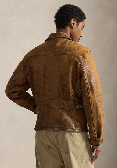 Καφέ δερμάτινο μπουφάν με υφή, ζωνάρι στη μέση και μυτερό γιακά. Διαθέτει ραφές στους ώμους και μακριά μανίκια.