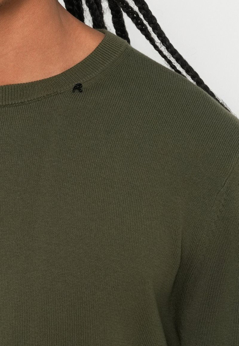 Dunkelgrüner Strickpullover mit einem gerippten Rundhalsausschnitt und schwarzem Akzentstich nahe dem Kragen. Die Textur ist weich und gleichmäßig.