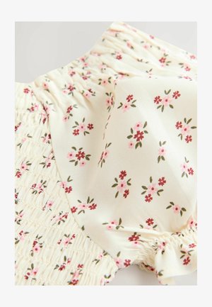 Blommönstrad tyg med små rosa och röda blommor på en krämfärgad bakgrund, med en smockad textur längs fållen.