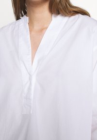 Vit bomullstunika med v-ringning, subtil textur och en lös, avslappnad passform. Ärmarna är långa och något avsmalnande.