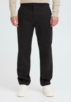 Mężczyzna w czarnych, casualowych spodniach, beżowym swetrze i białych trampkach, stojący na tle jasnego, jednolitego tła.