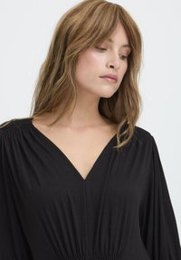 Rochie neagră cu decolteu în V, talie elastică și mâneci lungi, voluminoase. Textură netedă și design simplu. Fundal neutru.
