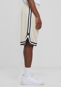 Vue de profil d'une personne portant un short de basketball beige avec des rayures noires et blanches, des baskets blanches et des chaussettes sur fond uni.