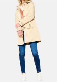 Béžový nepromokavý trenčkot s kapucí, zapínáním na knoflíky vpředu a páskem v pase; v kombinaci s modrými úzkými džínami a bílými teniskami.