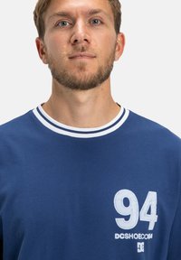 Modrá tričko s okrúhlym výstrihom a bielym prúžkovaným golierom, so štýlovým veľkým bielym potlačou "94" a logom. Vyrobené z textúrovaného materiálu, navrhnuté pre pohodlie.