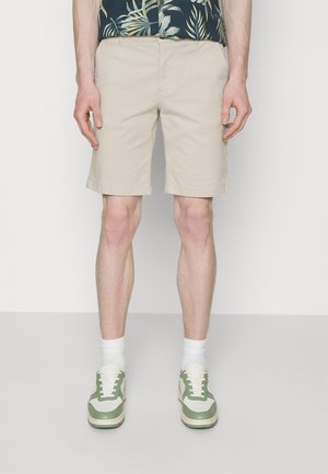 Homme portant un short beige jusqu’aux genoux, des chaussettes blanches et des baskets blanches et vertes, debout devant un fond clair uni.