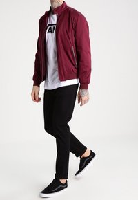 Burgundowa kurtka bomber z wysokim kołnierzem, z kieszeniami na zamek, noszona na białym t-shircie z grafiką, w zestawie z czarnymi spodniami i czarnymi sneakersami.