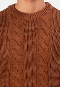Primo piano di un petto che indossa un maglione di lana marrone con motivi a treccia e un collo a coste.