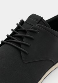 Zapato negro con cordones que presenta un material superior texturizado, cordones redondos y una suela blanca contrastante con un detalle mínimo.