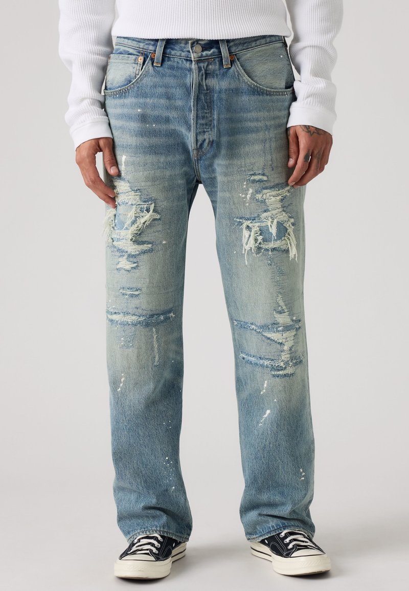 Homme portant un jean bleu clair déniché avec des patchs déchirés et des éclaboussures de peinture blanche, une chemise blanche à manches longues, et des baskets basses noires.