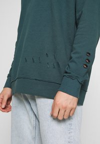 Felpa a maniche lunghe color teal con dettagli invecchiati e accenti filati. Maniche con polsini e piccoli tagli. Materiale in cotone morbido.