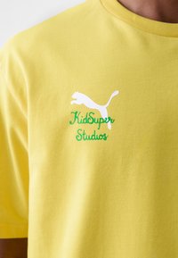 Camiseta de algodón amarilla con el logo blanco de Puma y el texto verde "KidSuper Studios", cuello redondo y mangas cortas, textura suave.