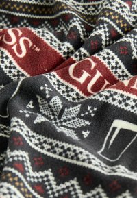 Koc polarowy w czarno-biało-bordowe wzory, z płatkami śniegu i logo "GUESS" w wyrazistym czerwonym kolorze. Miękka, teksturowana tkanina.