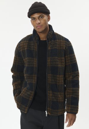 Mann trägt eine braune Strickmütze, eine fleece Jacke in Marineblau und Braun mit Karomuster, einen schwarzen Pullover und dunkle Hose, steht mit teilweise in die Taschen gesteckten Händen.
