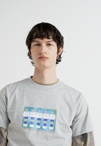 Šedé bavlnené tričko s potlačou piatich postáv v športových uniformách. Dizajn zahŕňa farebné akcenty a hladkú textúru.
