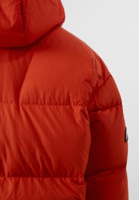 Rød puffjakke med quiltet design, hette og en strukturert overflate; har kontraststing og et logo-patch på siden.