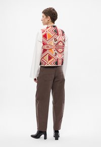 Színes, geometrikus mintával hímzett mellény piros, rózsaszín és fekete színekben. Fehér ing felett viselve, barna nadrággal és fekete csizmával párosítva.