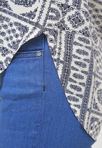 Weißes Hemd mit schwarzem Paisleymuster, kombiniert mit blauen Jeans. Das Hemd hat einen seitlichen Schlitz; die Jeans haben eine vordere Tasche mit einem Knopfdetail.