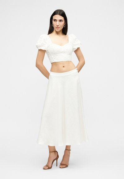 Femme aux longs cheveux foncés portant un crop top blanc à manches bouffantes et une jupe midi blanche avec des sandales à talons marron, debout, les mains dans le dos.