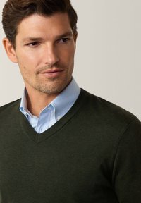 Suéter de punto en verde oscuro con un diseño texturizado en cuello en V, superpuesto a una camisa de cuello azul claro. Vista en primer plano del escote y los detalles de la tela.