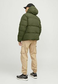 Olivengrøn pufferjakke med hætte, ribbede manchetter og en kort pasform. Bæres over beige cargo-bukser og sorte sportsko.