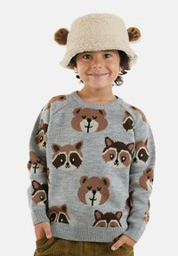 Ein Kind trägt einen beigen, flauschigen Bucket Hat mit Bärenohren und einen grauen Pullover mit Waschbären- und Bärenmotiven. Es lächelt und hat die Hände in den Taschen.