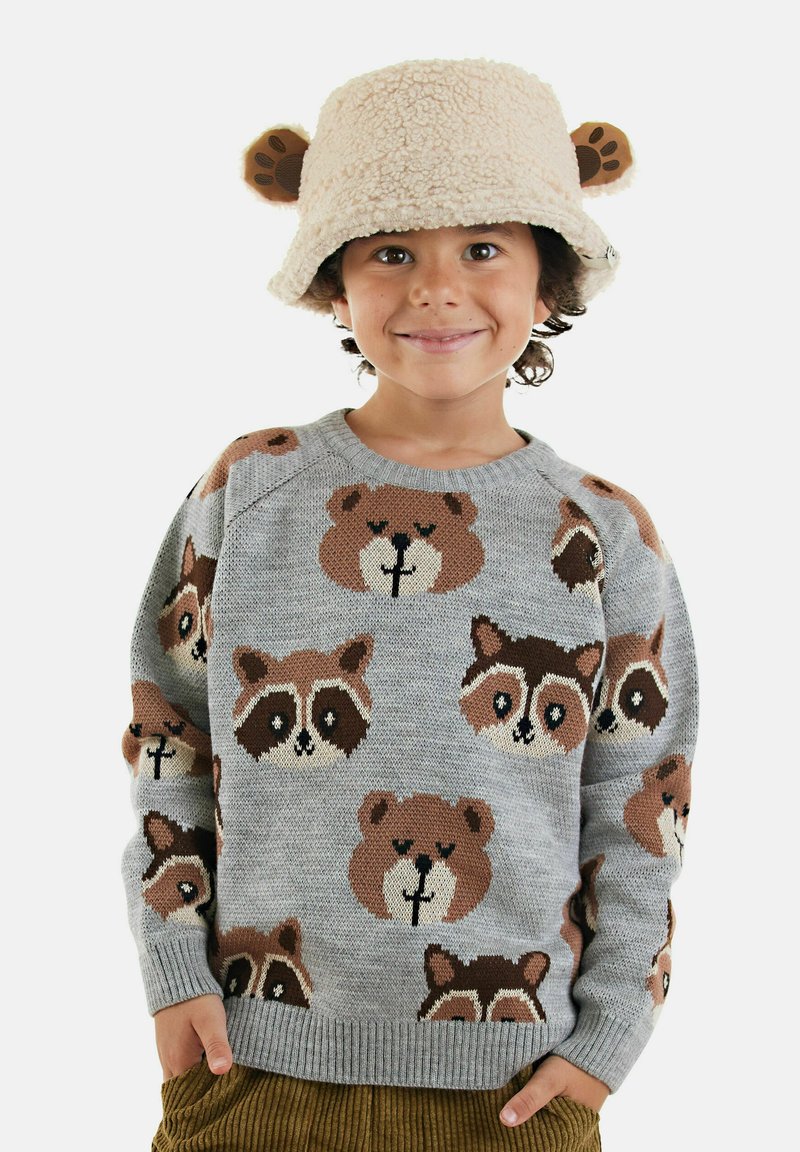 Ein Kind trägt einen beigen, flauschigen Bucket Hat mit Bärenohren und einen grauen Pullover mit Waschbären- und Bärenmotiven. Es lächelt und hat die Hände in den Taschen.