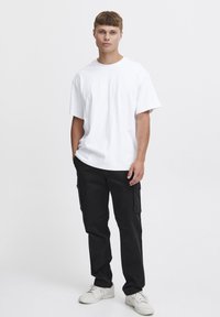 Weißes Baumwoll-T-Shirt mit lockerem Schnitt, kombiniert mit schwarzen Cargo-Hosenträgern mit Seitentaschen und weißen Sneakers. Einfaches, lässiges Outfit.