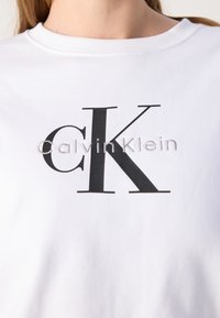T-shirt en coton blanc avec un grand logo "CK" noir et le texte "CalvinKlein" brodé en gris. Encolure ronde et texture lisse.